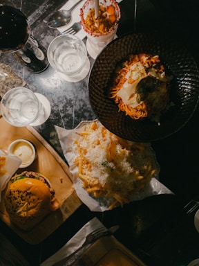 A table set with various dishes including a burger served on a wooden tray with a side of sauce, a plate of pasta with cheese and herbs, a pile of fries topped with shredded cheese, and a dessert or milkshake garnished with strawberries. Two glasses of water and a glass of red wine accompany the meal.