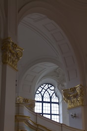 An ornate interior with a high ceiling featuring intricate sculptural details and embellishments. A large arched window with decorative metalwork on its frame allows natural light to illuminate the space. The architectural style is highlighted by elegant, gilded moldings and Corinthian columns.