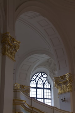 An ornate interior with a high ceiling featuring intricate sculptural details and embellishments. A large arched window with decorative metalwork on its frame allows natural light to illuminate the space. The architectural style is highlighted by elegant, gilded moldings and Corinthian columns.