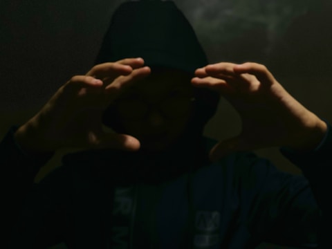 A person wearing a dark hoodie is partially obscured in shadows, with their hands raised and forming a frame around their face. The lighting is low and creates a mysterious atmosphere.