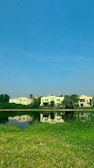 A tranquil scene of a lakeside with several modern houses in the background, surrounded by lush greenery. The clear, blue sky reflects in the calm water, creating a serene atmosphere with a hint of nature's beauty.