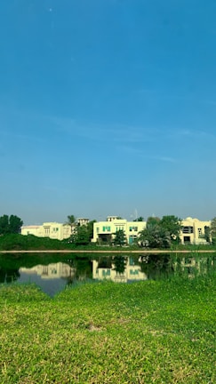A tranquil scene of a lakeside with several modern houses in the background, surrounded by lush greenery. The clear, blue sky reflects in the calm water, creating a serene atmosphere with a hint of nature's beauty.