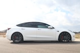 A sleek, modern electric car is parked on a paved road, with a clear view of its aerodynamic design and stylish appearance. The car is white with tinted windows, and it features large black wheels with red brake calipers. The surrounding area is a suburban neighborhood with some visible houses in the background, under a partly cloudy sky.