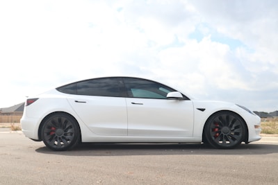 A sleek, modern electric car is parked on a paved road, with a clear view of its aerodynamic design and stylish appearance. The car is white with tinted windows, and it features large black wheels with red brake calipers. The surrounding area is a suburban neighborhood with some visible houses in the background, under a partly cloudy sky.