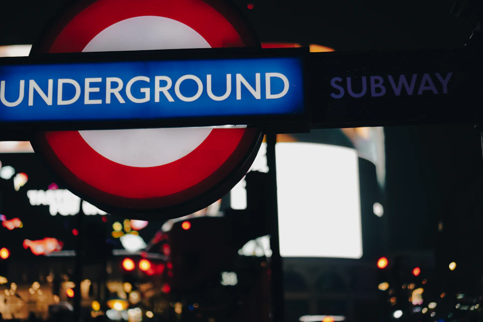 The image shows a close-up view of a subway station sign, which is predominantly red and white with a blue "Underground" logo at its center. The sign is mounted on a pole that extends into the background, providing information about the subway system to commuters. In the blurred background, there are other signs and advertisements for various businesses, indicating the busy nature of the area around the subway station.