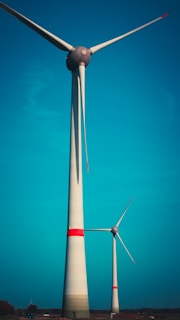 Two large wind turbines stand against a bright blue sky. The turbines have long, sleek blades and are prominently highlighted by horizontal red stripes near the top of their towers. They are situated on a flat landscape with sparse vegetation visible at the base.