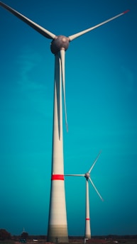 Two large wind turbines stand against a bright blue sky. The turbines have long, sleek blades and are prominently highlighted by horizontal red stripes near the top of their towers. They are situated on a flat landscape with sparse vegetation visible at the base.