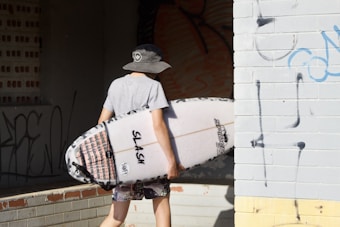 A person carrying a surfboard walks towards a building with graffiti-covered walls. The person is wearing a wide-brimmed hat, a gray t-shirt, and patterned shorts. The surfboard has a design and some text on it.