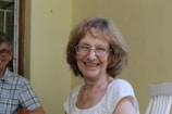 An elderly woman with glasses is smiling warmly, seated indoors. She wears a white blouse and appears to be enjoying herself. A man wearing glasses and a plaid shirt is partly visible, sitting next to her.