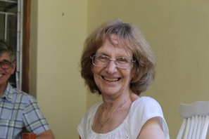 An elderly woman with glasses is smiling warmly, seated indoors. She wears a white blouse and appears to be enjoying herself. A man wearing glasses and a plaid shirt is partly visible, sitting next to her.