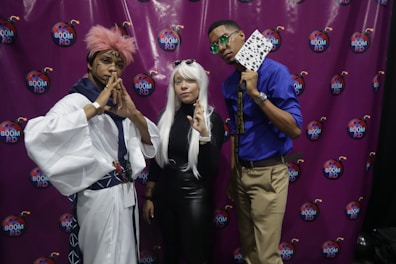 Three individuals pose prominently in cosplay attire against a purple backdrop featuring 'K Boom RD' logos. The left person wears a white outfit with pink hair and a traditional Japanese style headband. The middle person is dressed in black with long white hair and round sunglasses resting on their head. The right person wears a blue shirt, beige pants, and sunglasses, holding playing cards. Their poses suggest a playful and confident theme.