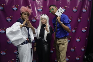 Three individuals pose prominently in cosplay attire against a purple backdrop featuring 'K Boom RD' logos. The left person wears a white outfit with pink hair and a traditional Japanese style headband. The middle person is dressed in black with long white hair and round sunglasses resting on their head. The right person wears a blue shirt, beige pants, and sunglasses, holding playing cards. Their poses suggest a playful and confident theme.
