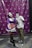 Two people are wearing unique masks. One is dressed in a purple outfit with a white ruffled apron and holding a prop weapon. The other is in casual clothing with camouflage pants and a printed t-shirt. They are posing playfully in front of a photo booth backdrop featuring a repeated circular logo.