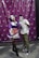 Two people are wearing unique masks. One is dressed in a purple outfit with a white ruffled apron and holding a prop weapon. The other is in casual clothing with camouflage pants and a printed t-shirt. They are posing playfully in front of a photo booth backdrop featuring a repeated circular logo.