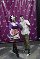Two people are wearing unique masks. One is dressed in a purple outfit with a white ruffled apron and holding a prop weapon. The other is in casual clothing with camouflage pants and a printed t-shirt. They are posing playfully in front of a photo booth backdrop featuring a repeated circular logo.