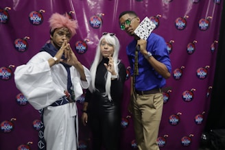 Three individuals dressed in elaborate costumes pose in front of a purple backdrop with 'Boom RD' logos. One person wears a white and blue robe with a pink wig, making a gesture with their hands. The person in the middle has long white hair and is wearing sunglasses and a form-fitting black outfit, also making a gesture with their hand. The third person wears a blue shirt, khaki pants, and holds a fan with a black and white pattern.