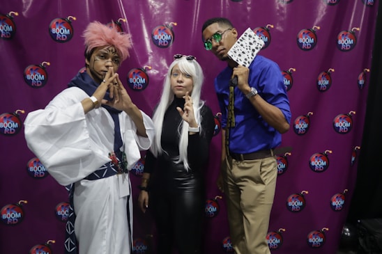 Three individuals dressed in elaborate costumes pose in front of a purple backdrop with 'Boom RD' logos. One person wears a white and blue robe with a pink wig, making a gesture with their hands. The person in the middle has long white hair and is wearing sunglasses and a form-fitting black outfit, also making a gesture with their hand. The third person wears a blue shirt, khaki pants, and holds a fan with a black and white pattern.