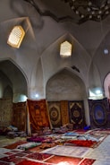 An interior scene featuring a collection of richly decorated carpets displayed against walls and laid on the floor. The room has arched ceilings with small, intricate windows allowing soft, diffused light to enter. Various carpets with ornate patterns and a range of colors are stacked and spread across a tiled floor, creating a vibrant display.