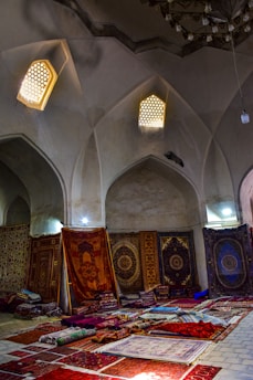 An interior scene featuring a collection of richly decorated carpets displayed against walls and laid on the floor. The room has arched ceilings with small, intricate windows allowing soft, diffused light to enter. Various carpets with ornate patterns and a range of colors are stacked and spread across a tiled floor, creating a vibrant display.