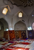 An interior scene featuring a collection of richly decorated carpets displayed against walls and laid on the floor. The room has arched ceilings with small, intricate windows allowing soft, diffused light to enter. Various carpets with ornate patterns and a range of colors are stacked and spread across a tiled floor, creating a vibrant display.