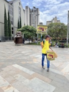 A person in a yellow shirt and blue jeans is holding a sign with an image of an ant and the words 'HORMIGA CULONA'. They are in a plaza area with a large bronze statue of a seated figure nearby. Tall buildings and greenery, including trees and bushes, are present in the background. A partly cloudy sky is visible above.