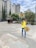 A person in a yellow shirt and blue jeans is holding a sign with an image of an ant and the words 'HORMIGA CULONA'. They are in a plaza area with a large bronze statue of a seated figure nearby. Tall buildings and greenery, including trees and bushes, are present in the background. A partly cloudy sky is visible above.
