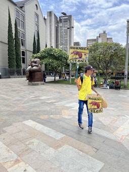 A person in a yellow shirt and blue jeans is holding a sign with an image of an ant and the words 'HORMIGA CULONA'. They are in a plaza area with a large bronze statue of a seated figure nearby. Tall buildings and greenery, including trees and bushes, are present in the background. A partly cloudy sky is visible above.