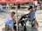 In a public square, a shoe shiner works diligently on a customer's shoe, seated on a green plastic chair. They are under a red umbrella, surrounded by a few people sitting or walking in the background.