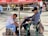 In a public square, a shoe shiner works diligently on a customer's shoe, seated on a green plastic chair. They are under a red umbrella, surrounded by a few people sitting or walking in the background.