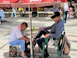 In a public square, a shoe shiner works diligently on a customer's shoe, seated on a green plastic chair. They are under a red umbrella, surrounded by a few people sitting or walking in the background.