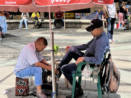 In a public square, a shoe shiner works diligently on a customer's shoe, seated on a green plastic chair. They are under a red umbrella, surrounded by a few people sitting or walking in the background.