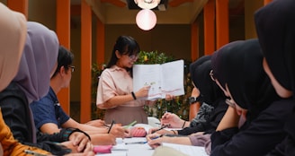 A group of people sits around a table, attentively looking at a woman who is holding and explaining contents from an open book. The setting appears to be indoors, with warm lighting and plants in the background, giving a formal yet relaxed atmosphere.