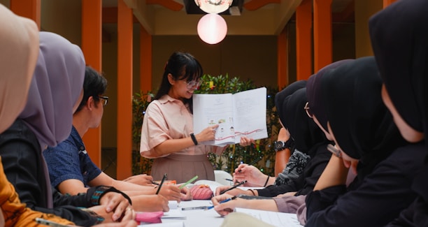 A group of people sits around a table, attentively looking at a woman who is holding and explaining contents from an open book. The setting appears to be indoors, with warm lighting and plants in the background, giving a formal yet relaxed atmosphere.