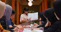 A group of people sits around a table, attentively looking at a woman who is holding and explaining contents from an open book. The setting appears to be indoors, with warm lighting and plants in the background, giving a formal yet relaxed atmosphere.