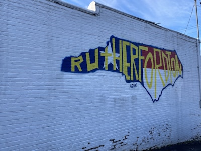 A large mural painted on a white brick wall depicts the outline of the state of North Carolina. Inside the outline, the word 'Rutherfordton' is written in bold yellow letters. The background within the state outline is colored with blue and red, with an artistic design. The wall shows patches of peeling paint, giving a rustic look.