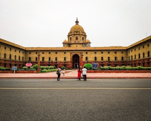 A large, symmetrical government building with a central dome, surrounded by a red-stone courtyard. Three people are standing in the foreground near a roadway, with trimmed green hedges visible by the building.