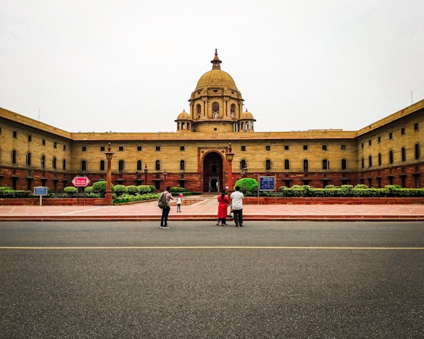 A large, symmetrical government building with a central dome, surrounded by a red-stone courtyard. Three people are standing in the foreground near a roadway, with trimmed green hedges visible by the building.