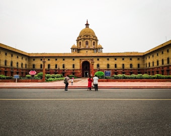 A large, symmetrical government building with a central dome, surrounded by a red-stone courtyard. Three people are standing in the foreground near a roadway, with trimmed green hedges visible by the building.