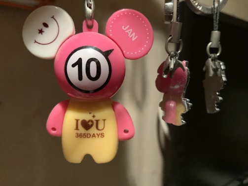A small, colorful keychain shaped like a bear with cartoonish features, including pink ears, one with a smiley face and the other marked 'JAN'. The bear's body features a large '10' on its chest and a message that reads 'I ♥ U 365DAYS'. Several other keychains are visible to the right.