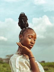 A person with intricately styled braids adorned with metallic clips, enhancing the elegance of their pose. Eyes are gently closed, with a serene expression on their face. The background features soft, cloudy skies adding a dreamy atmosphere.