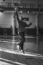 A striking black and white action shot of a basketball player mid-air, muscles tense, against a dark, blurred stadium background.