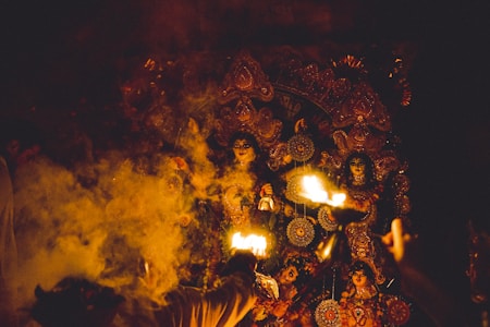The image captures a traditional religious ceremony featuring ornate, colorful decorations and idols. Smoke drifts through the scene, illuminated by bright flames in the foreground. The setting appears to be dimly lit, adding to the mystical and spiritual atmosphere.