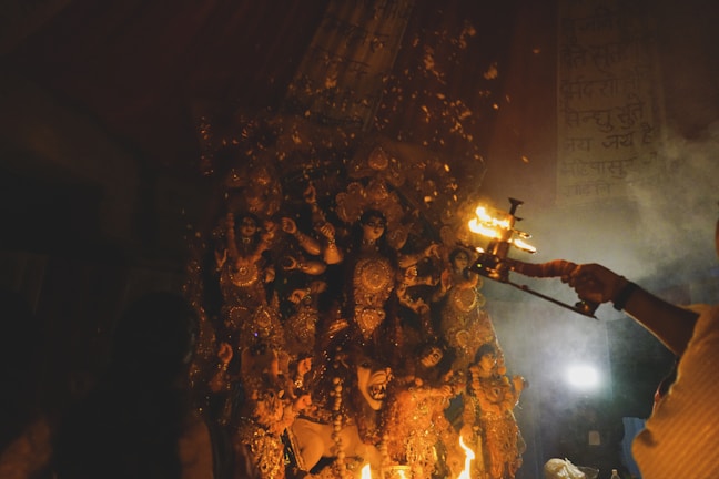 An elaborate statue of a multi-armed goddess is adorned with intricate decorations and surrounded by smaller figures, likely representing other deities or attendants. In the foreground, a person holds a lit ceremonial lamp, casting a warm glow and creating an atmosphere filled with smoke and reverence.