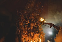 An elaborate statue of a multi-armed goddess is adorned with intricate decorations and surrounded by smaller figures, likely representing other deities or attendants. In the foreground, a person holds a lit ceremonial lamp, casting a warm glow and creating an atmosphere filled with smoke and reverence.