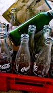 Empty glass soda bottles are arranged in a red plastic crate. The bottles have recognizable branding and are partially reflecting light. A green bottle is positioned horizontally on top of the crate. A stained paper is placed behind the bottles.