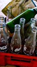 Empty glass soda bottles are arranged in a red plastic crate. The bottles have recognizable branding and are partially reflecting light. A green bottle is positioned horizontally on top of the crate. A stained paper is placed behind the bottles.
