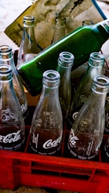 Empty glass soda bottles are arranged in a red plastic crate. The bottles have recognizable branding and are partially reflecting light. A green bottle is positioned horizontally on top of the crate. A stained paper is placed behind the bottles.