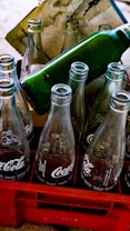 Empty glass soda bottles are arranged in a red plastic crate. The bottles have recognizable branding and are partially reflecting light. A green bottle is positioned horizontally on top of the crate. A stained paper is placed behind the bottles.