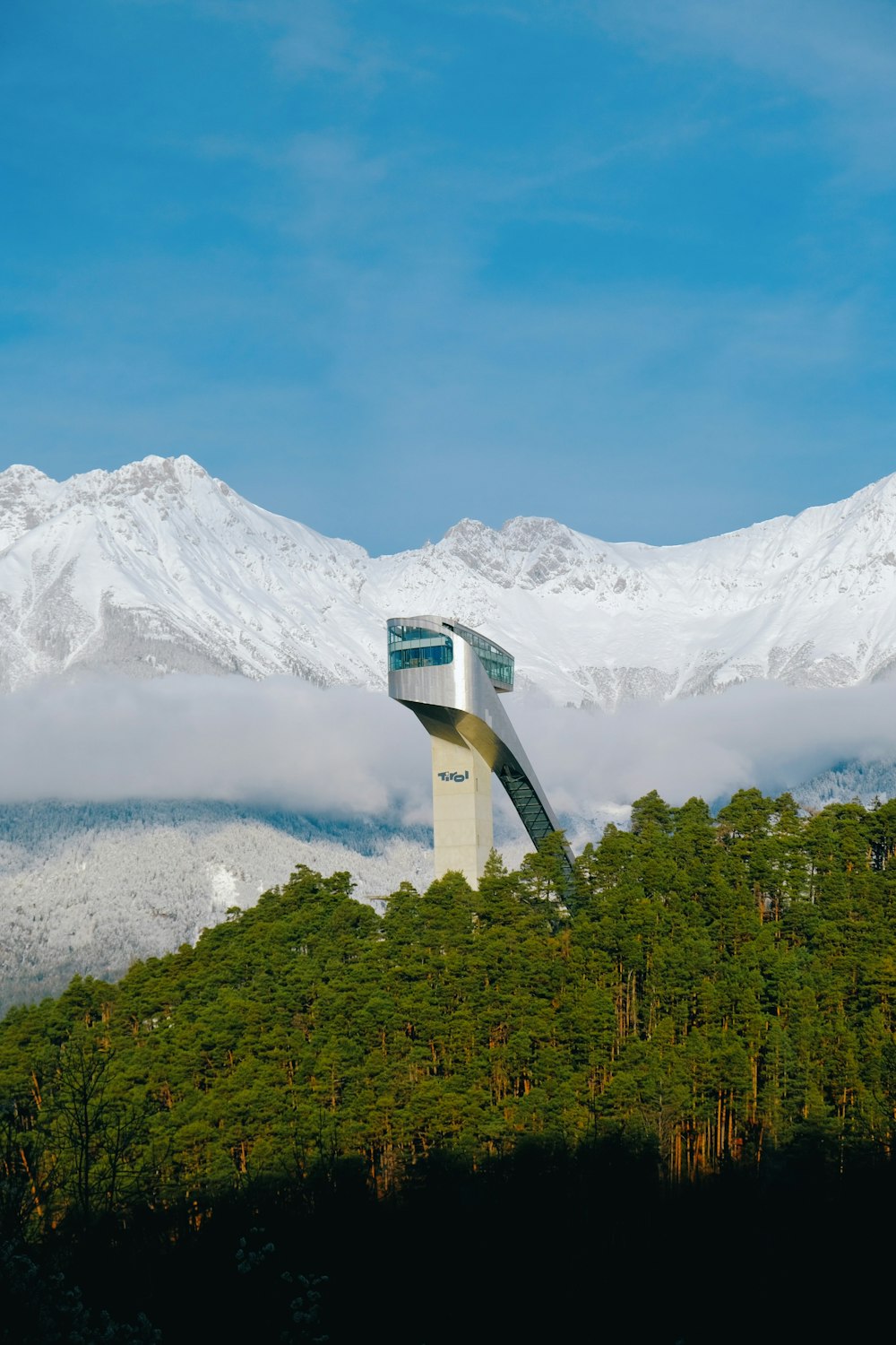 a tower in the middle of a forest with snow covered mountains in the background