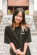 A young woman with black and light brown hair stands smiling in front of a stone staircase. She wears a black t-shirt and holds a small purse, with her hands intertwined. The background includes green foliage and part of a building with a tiled roof.